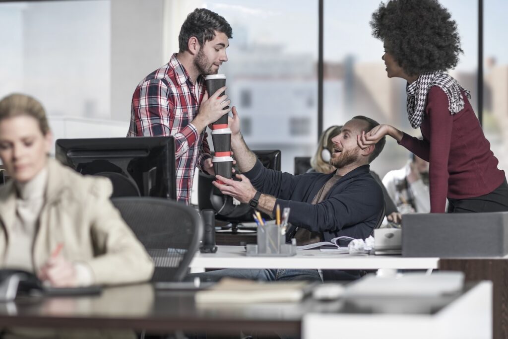 An example of a positive work environment where one employee helps a co-worker awkwardly handling 3 coffee cups.
